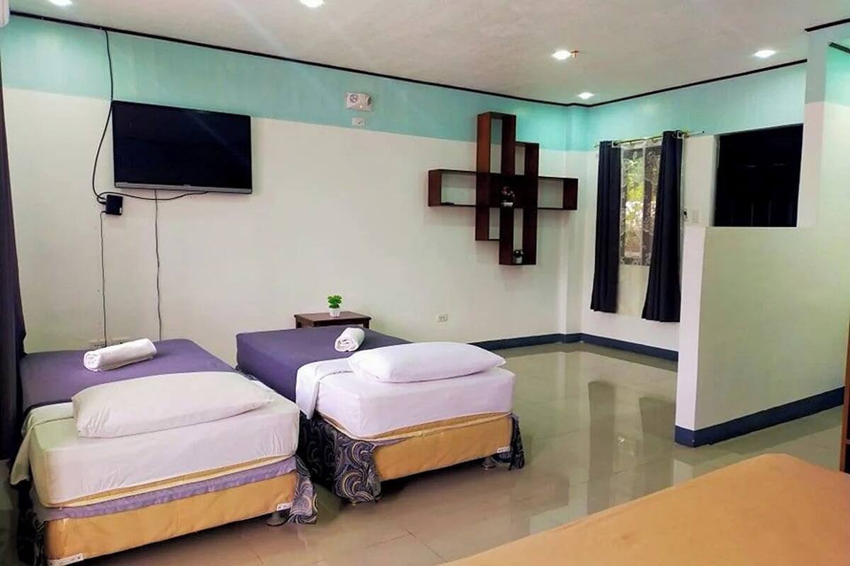 A hotel room with two beds, a TV mounted on the wall, a small table with a plant, and a cross-shaped decoration on the wall.
