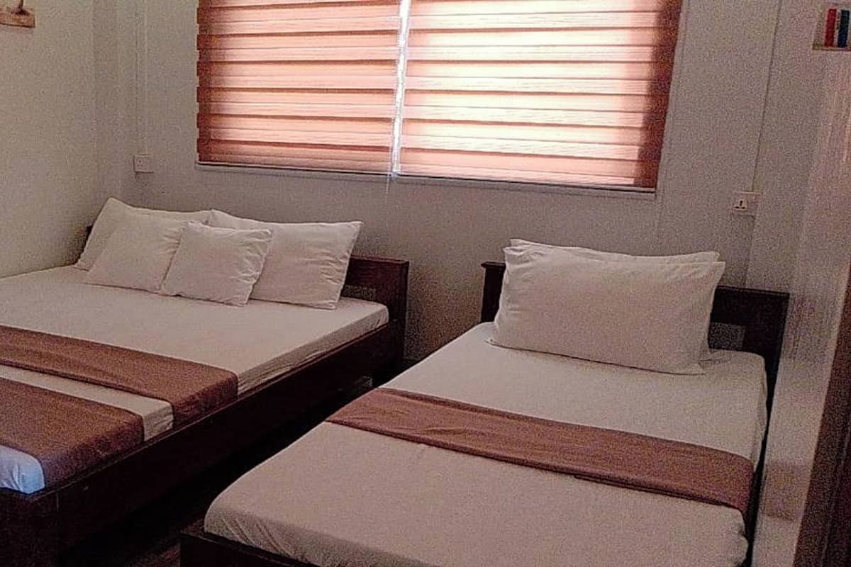 A room with two single beds, each with a wooden headboard and white bedding. There is a window with blinds, and the walls are painted a light color.