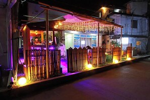 Un puesto de comida en la calle con una cerca de bambú, iluminado por luces de adorno y luces de neón.