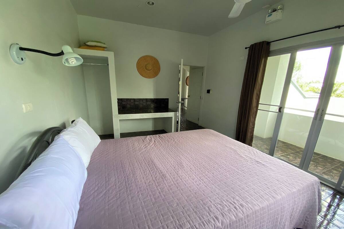Une chambre à coucher comprenant un lit, une table de chevet, une lampe fixée au mur, une porte donnant sur un balcon et un ventilateur de plafond.