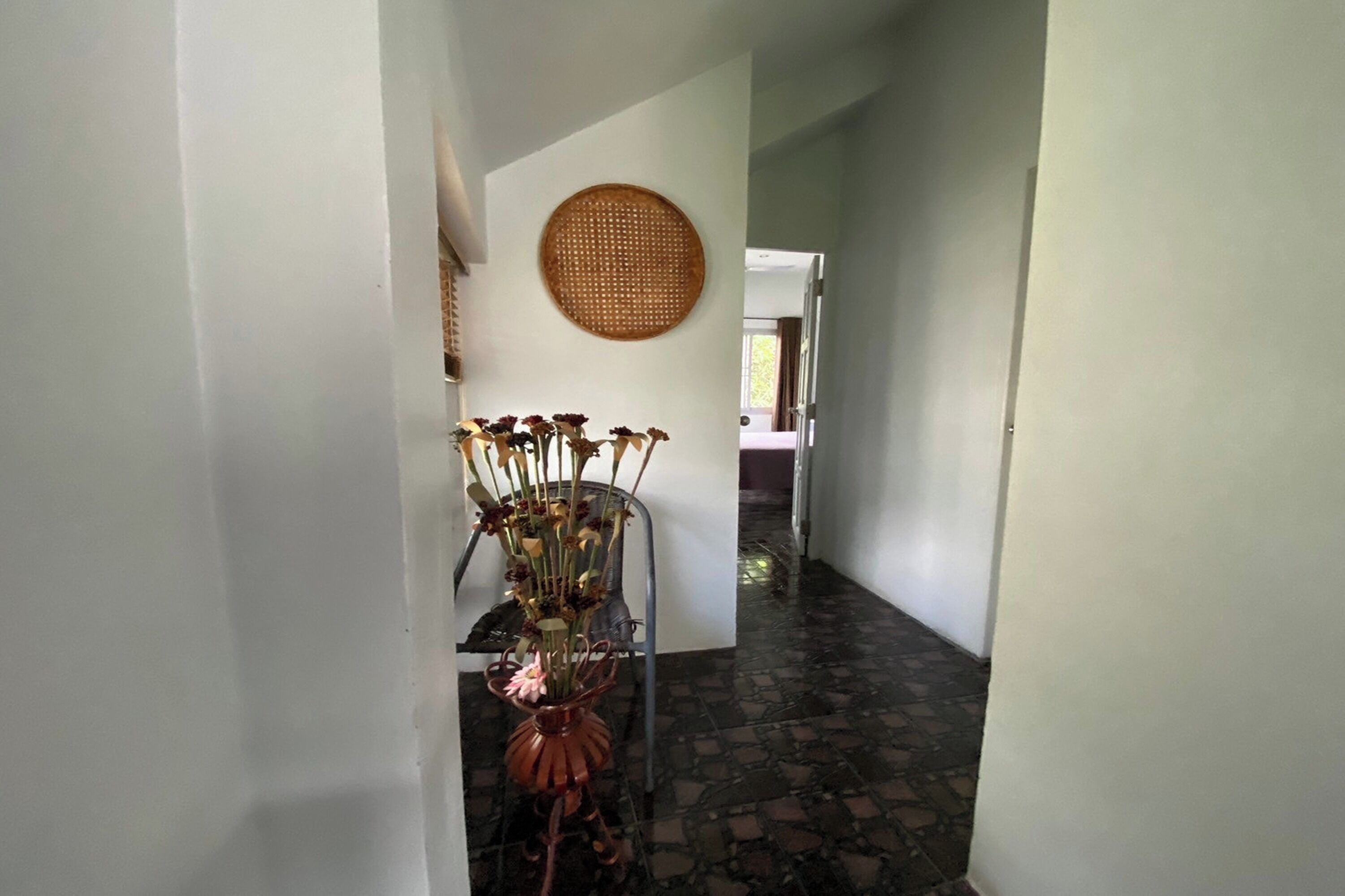 A hallway with a decorative vase on a metal stand, a woven wall hanging, and a tiled floor.