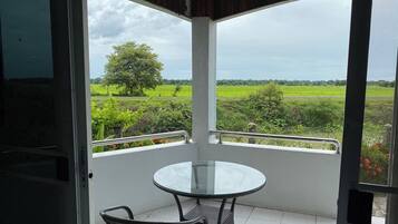 A balcony with a glass table and wicker chairs, overlooking a green landscape with trees and fields.