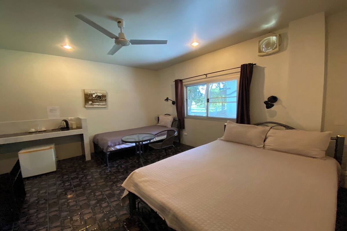 Une chambre d’hôtel comprenant un lit, un bureau, une chaise, une petite table, un ventilateur de plafond et une fenêtre avec des rideaux.