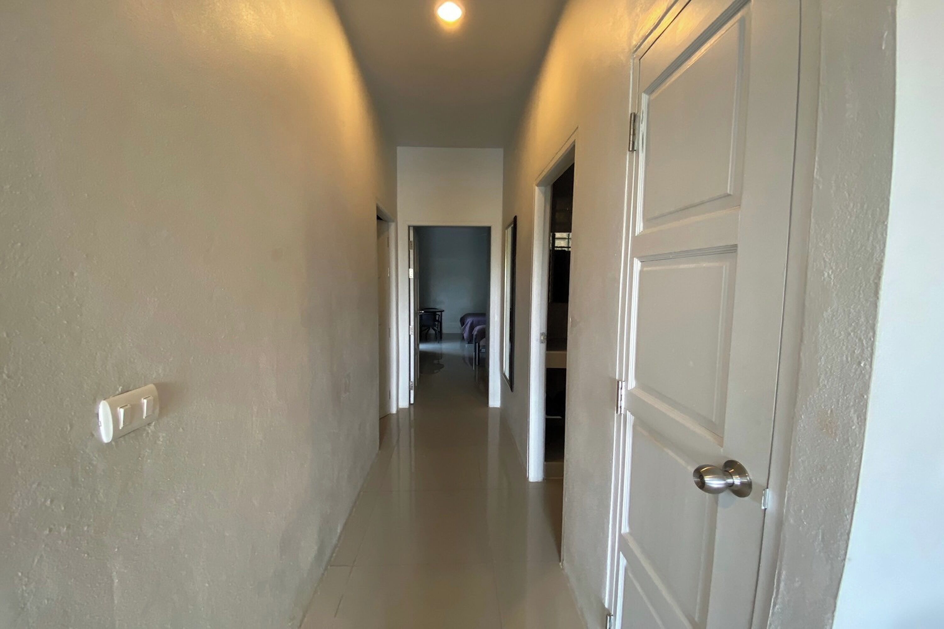 A hallway with white walls and doors, leading to a room with a bed and a chair.