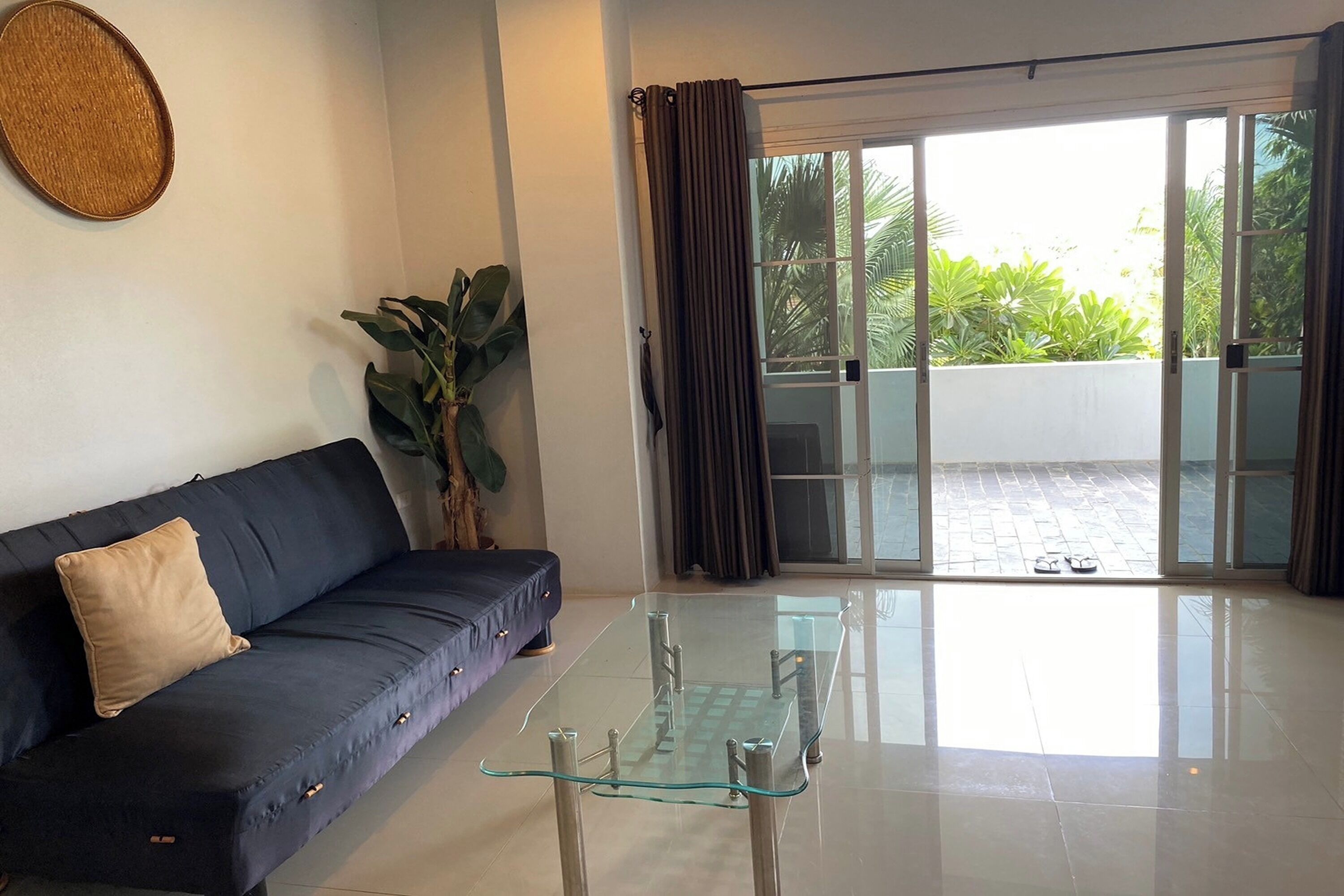 A living room with a dark blue sofa, a glass coffee table, a potted plant, and a view of a balcony with a tiled floor and greenery outside.