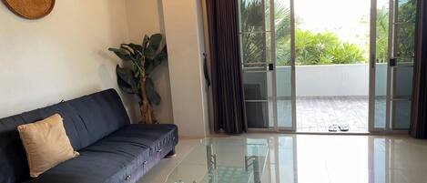 A living room with a dark blue sofa, a glass coffee table, a potted plant, and a view of a balcony with a tiled floor and greenery outside.