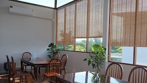 A dining area with a wooden table and chairs, large windows with blinds, and an air conditioning unit on the wall. - Burichan Resort (Chanthaburi)
