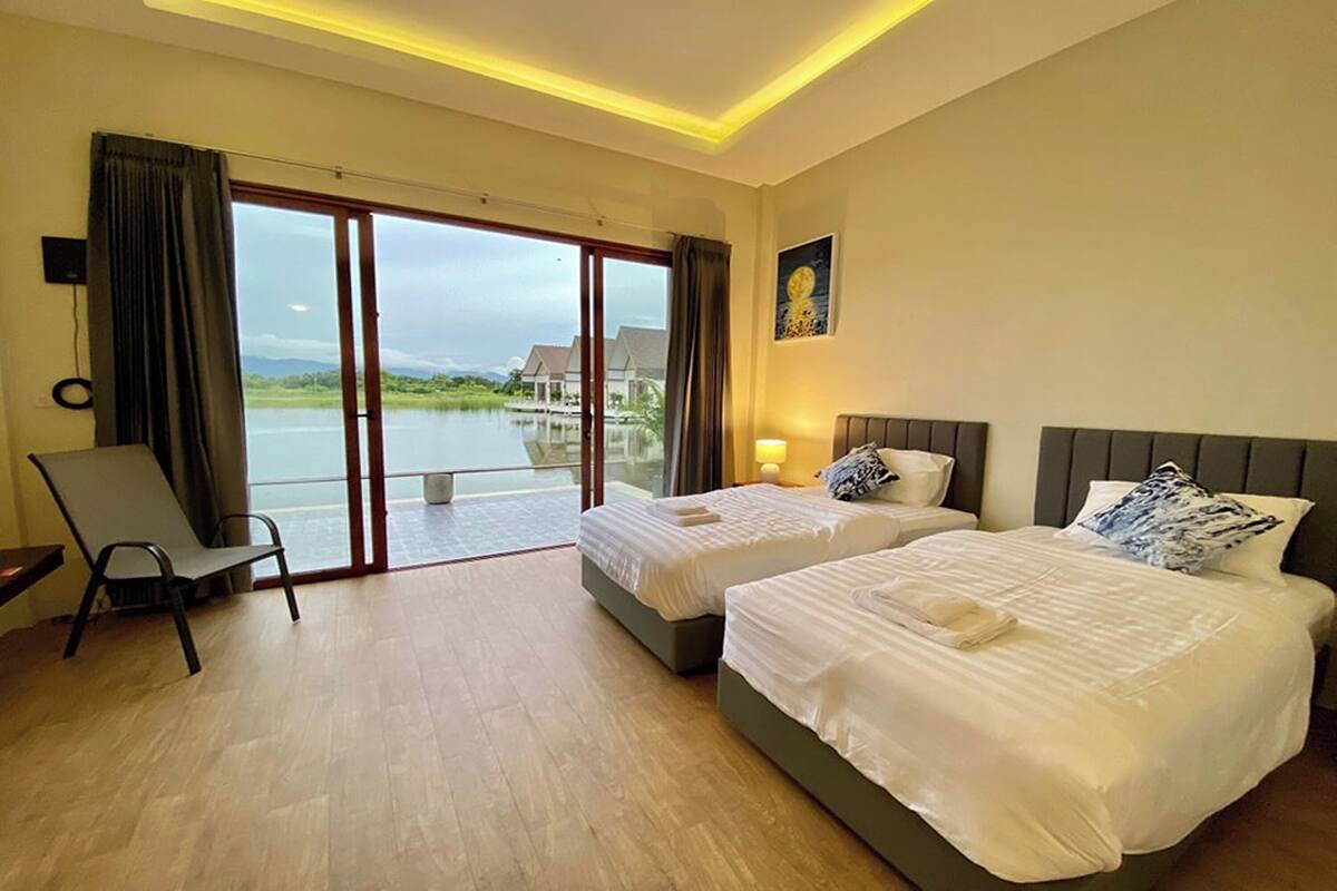 Ein Hotelzimmer mit zwei Betten, einem Sessel, einem großen Fenster mit Blick auf ein Gewässer und einem Bild an der Wand.