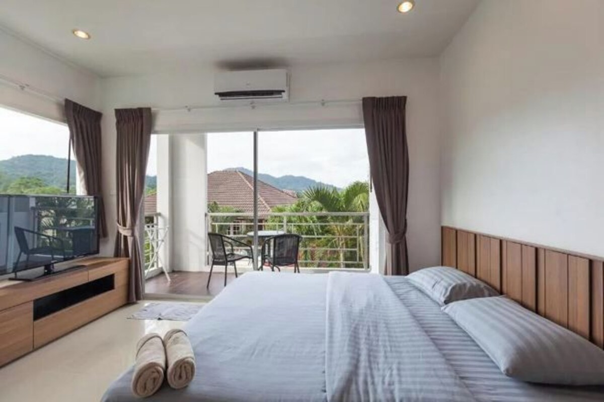 A hotel room with a bed, a TV, a balcony with chairs, and a view of the mountains.
