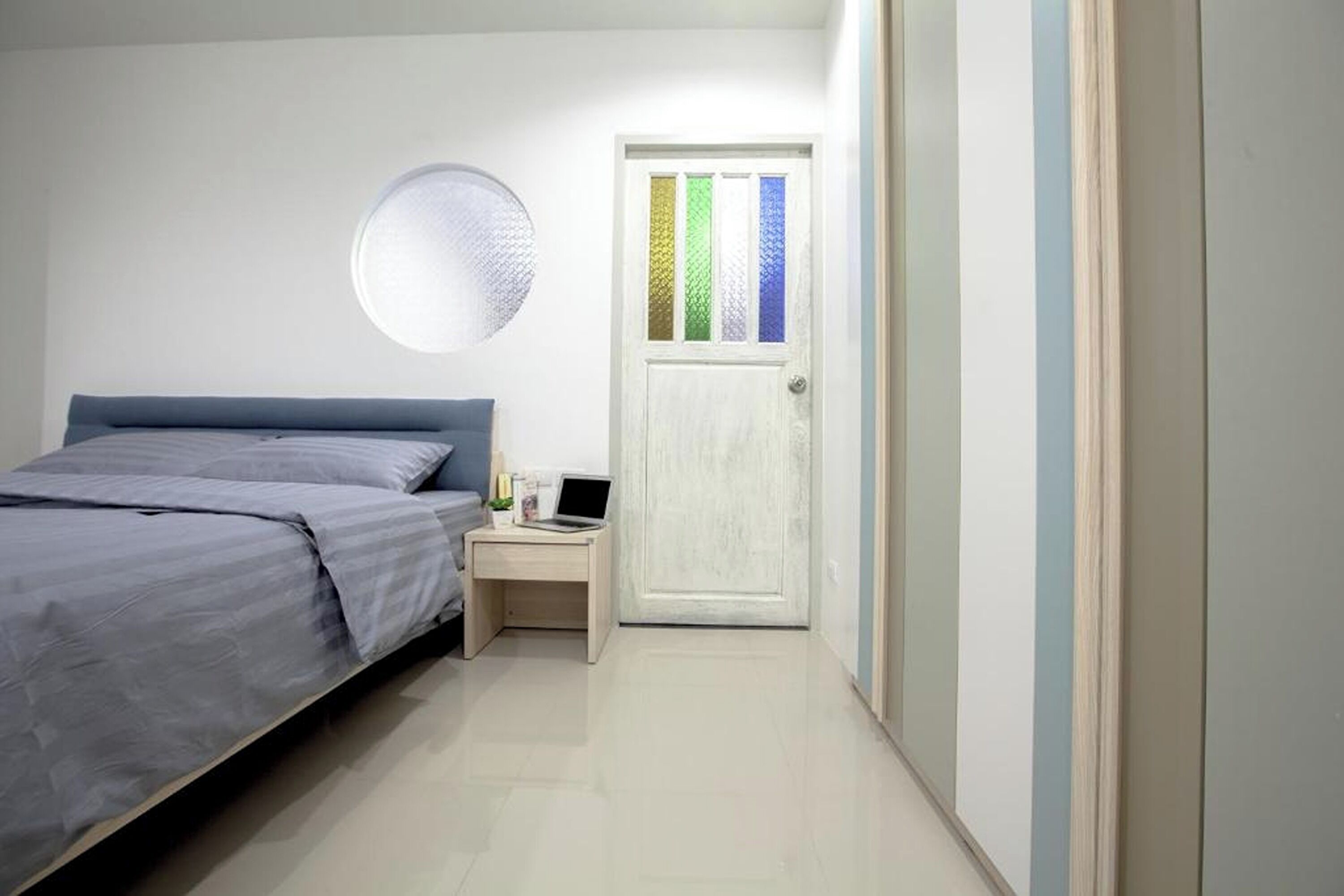 Quarto com cama, mesa de cabeceira, um laptop e porta com painéis de vidro coloridos.