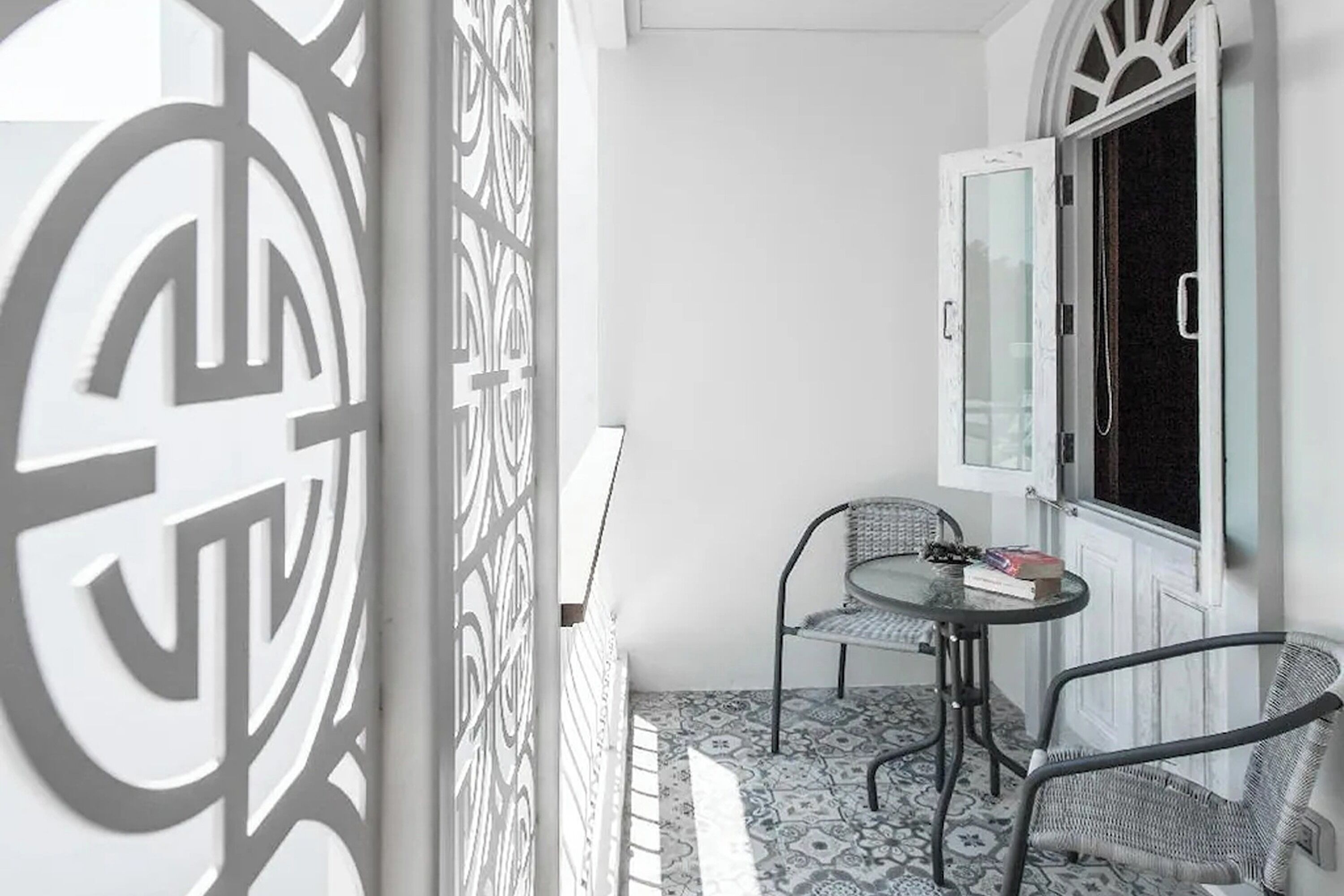 A small balcony with a patterned floor, two wicker chairs, a small table with a book, and a window with a curtain.