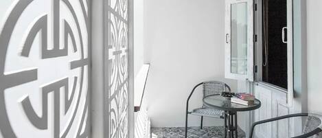 A small balcony with a patterned floor, two wicker chairs, a small table with a book, and a window with a curtain.
