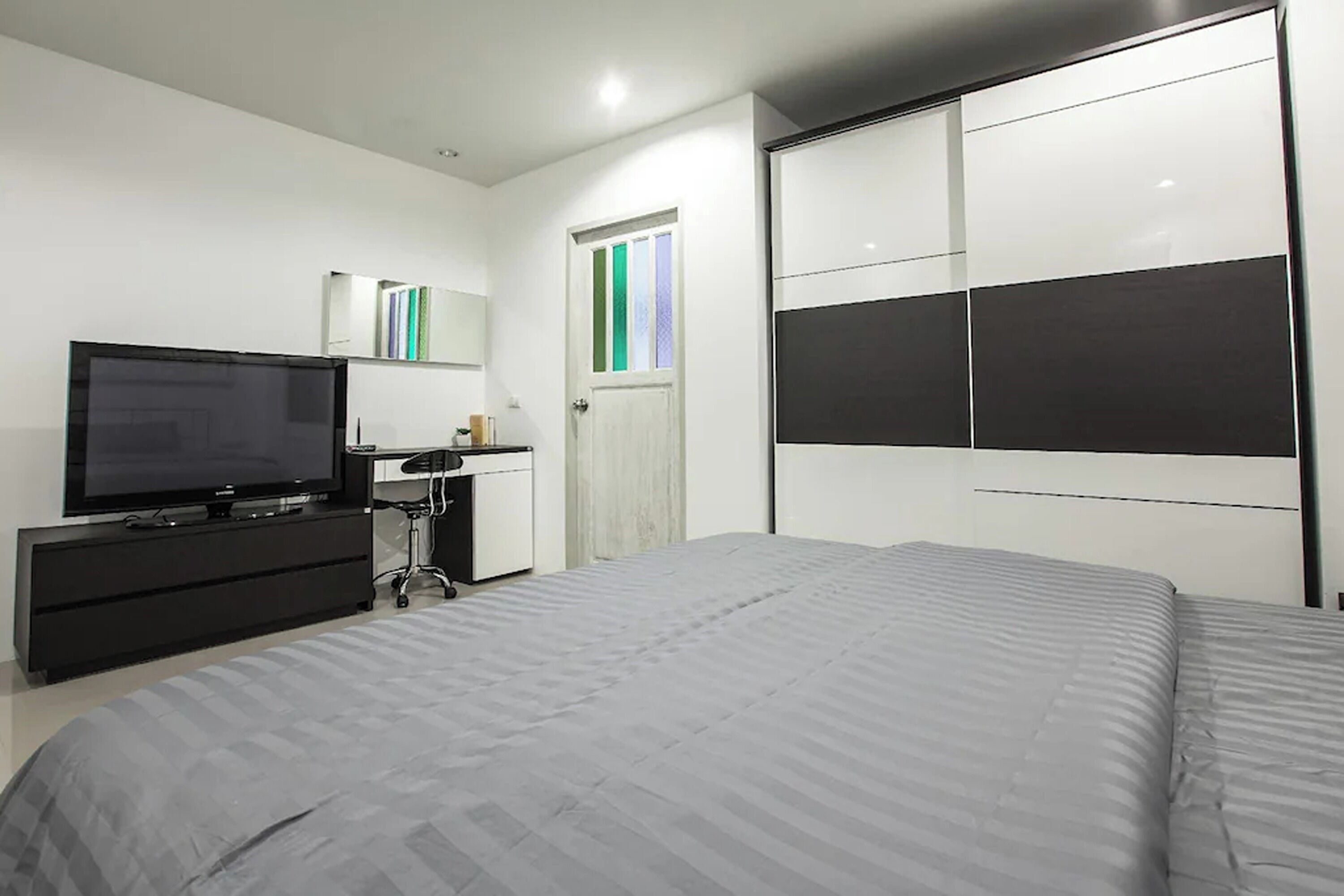 A modern bedroom with a large bed, a TV on a black dresser, a wardrobe with sliding doors, and a door with a green and white striped window.