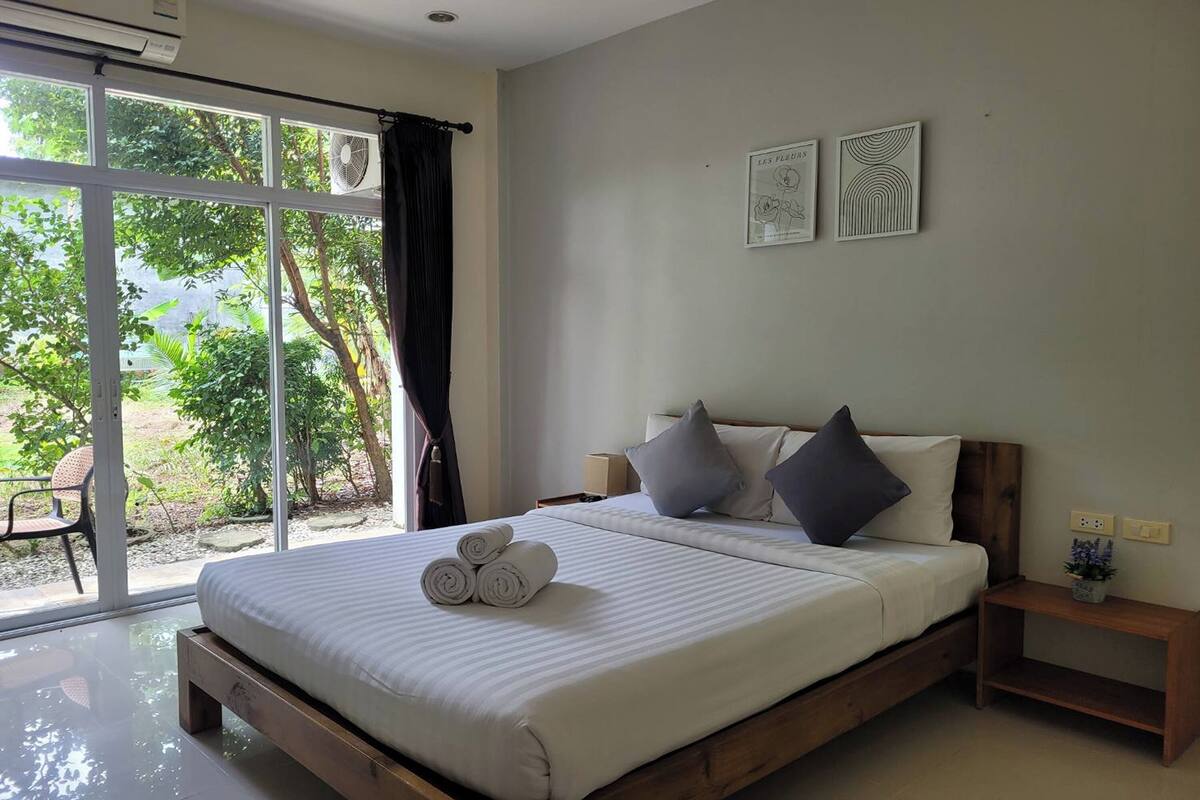 A bedroom with a large window, a bed with rolled towels, a bedside table, and a small nightstand.