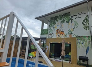 A pool area with a wooden deck, outdoor seating, and a mural on the building. - Win Win Villa (Sattahip)