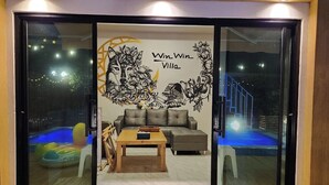 Un soggiorno con una porta vetrata che si apre su una zona piscina, un divano grigio, un tavolino in legno e un murale decorativo.