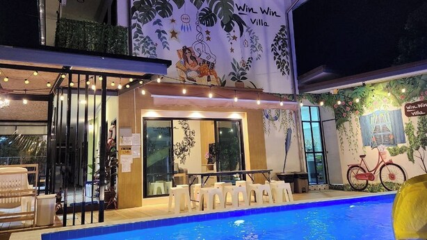 Zona piscina con area relax coperta, un murale raffigurante piante tropicali e una bicicletta, e uno striscione con la scritta "Win Win Villa".
