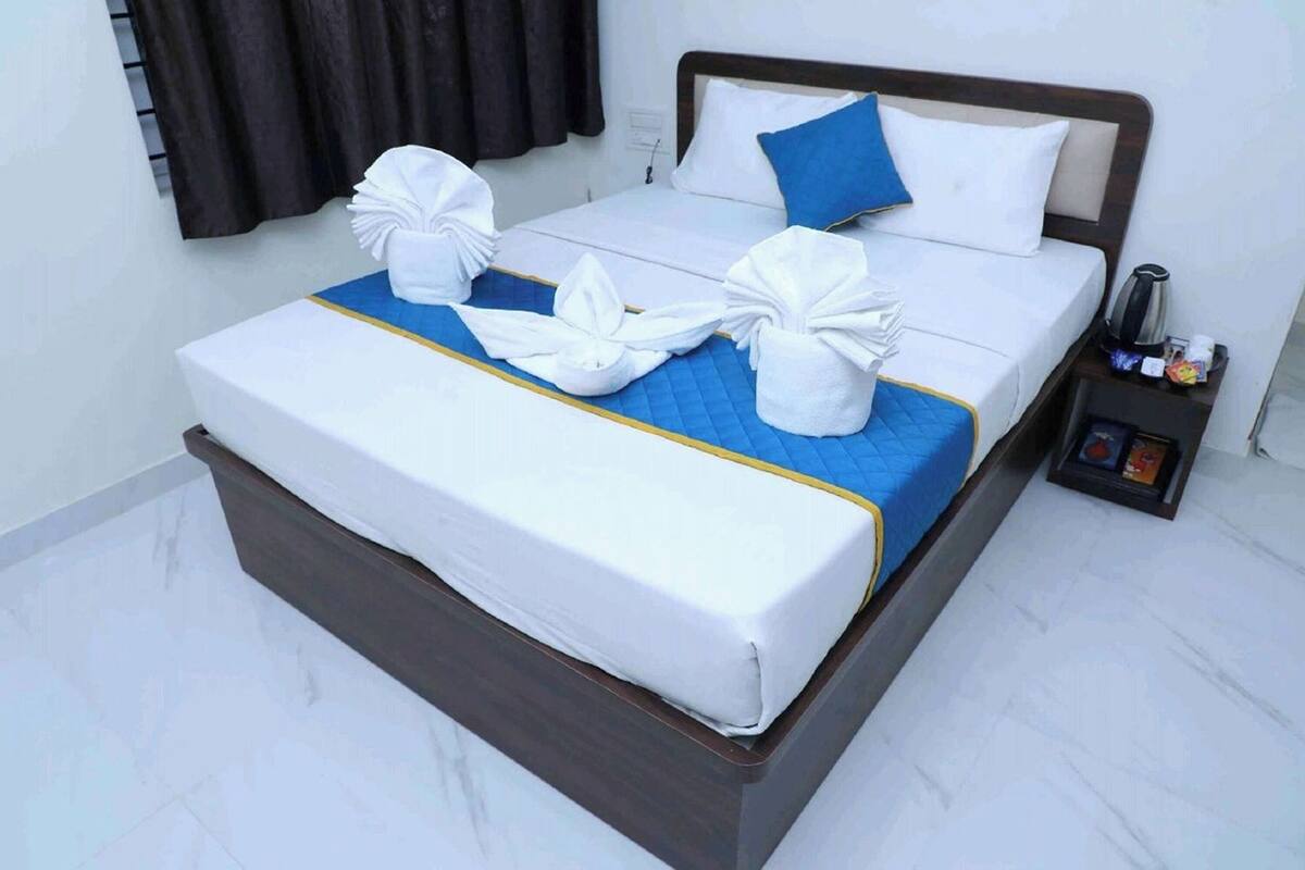 A neatly made bed with white linens and blue accents, a bedside table with a kettle and a small tray, and a dark wooden headboard.