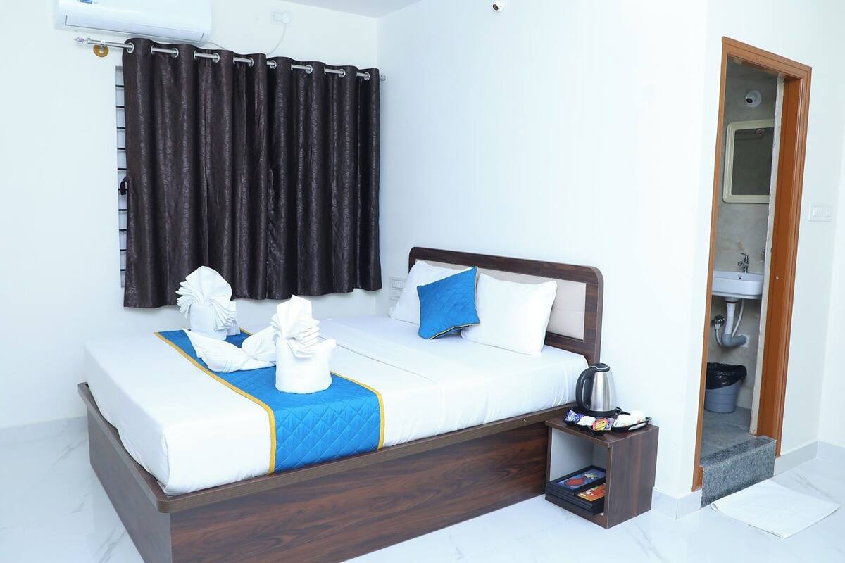 A neatly made bed with white linens and blue accents, a wooden headboard, a bedside table with a kettle and books, and a bathroom visible through an open door.