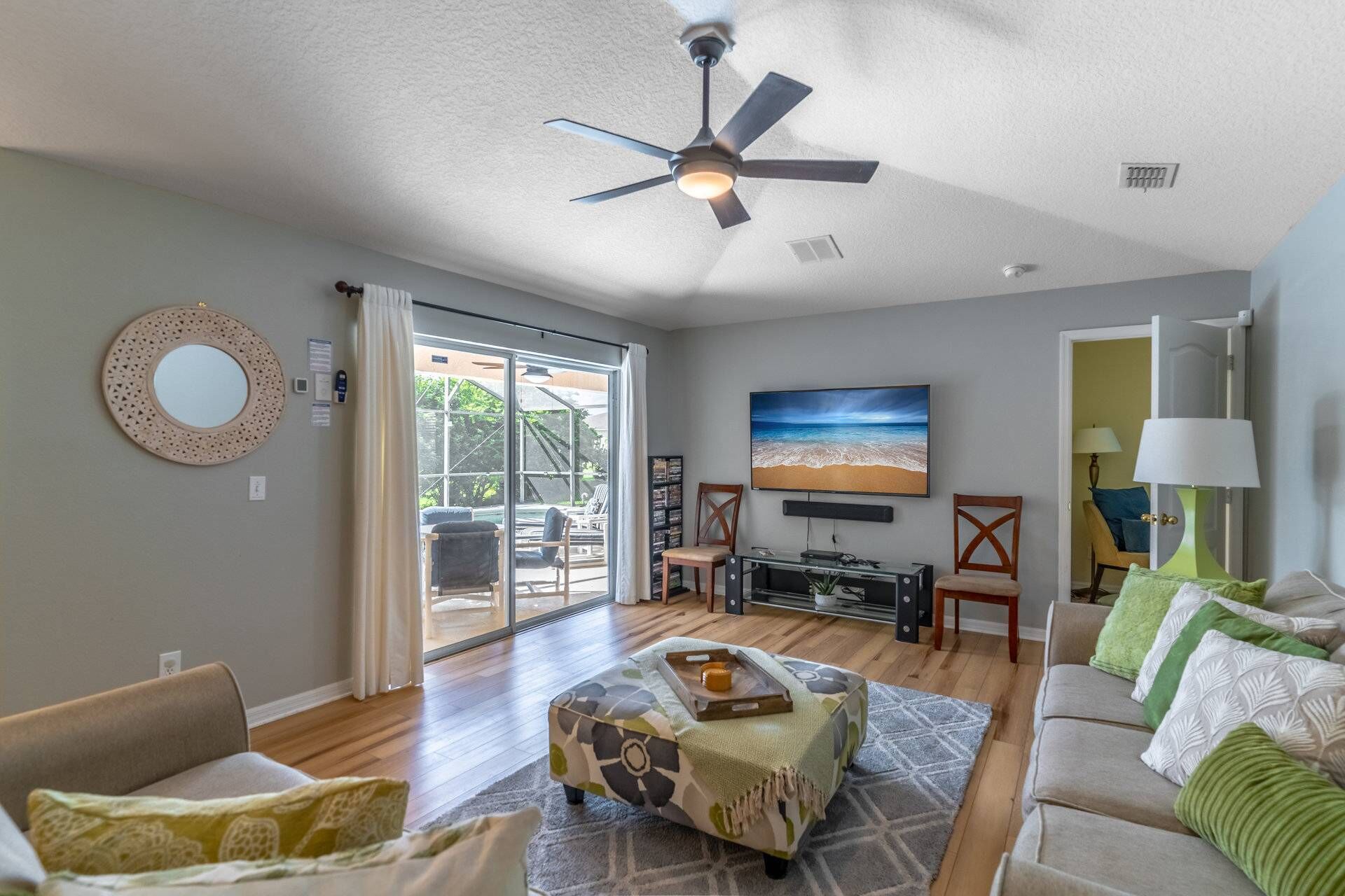 A spacious living room with a ceiling fan, a large TV mounted on the wall, and a comfortable seating area with a sofa and chairs.