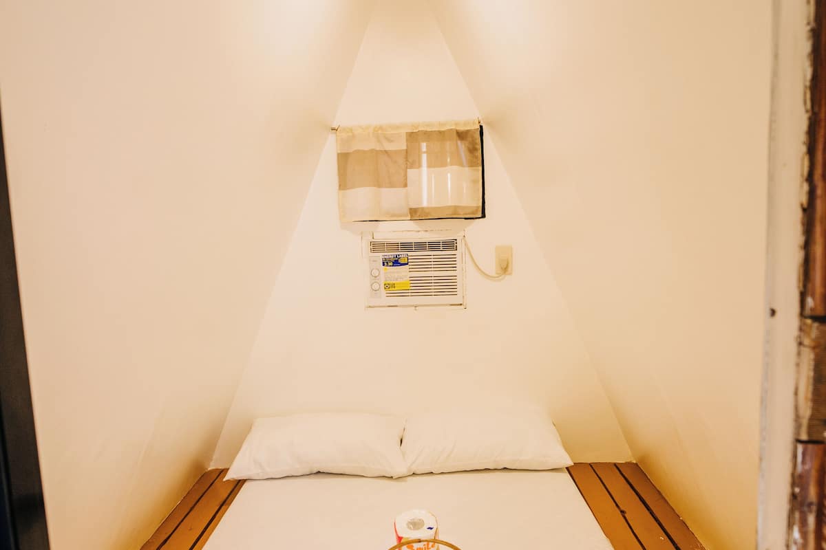 Una habitación pequeña y estrecha con una cama, una ventana con cortinas, un aire acondicionado instalado en la pared y una cesta con toallas en el suelo.