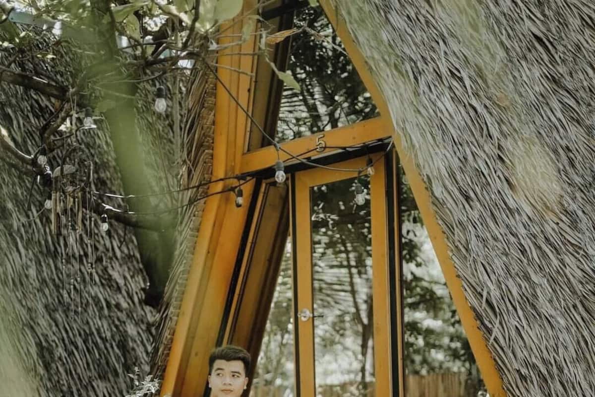 Una persona sentada en un banco de madera adosado a una estructura con techo de paja, rodeada de árboles.