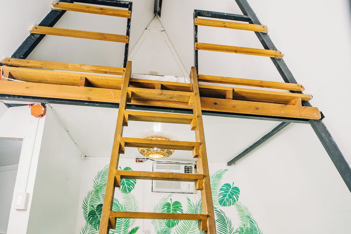 Una cama litera con escalera de madera, ventilador de techo y pared con papel tapiz de hojas tropicales.