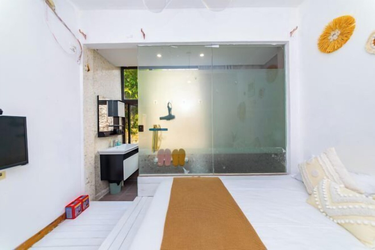 A modern bedroom with a large bed, a glass shower enclosure, a TV, and a bathroom with a sink and mirror.