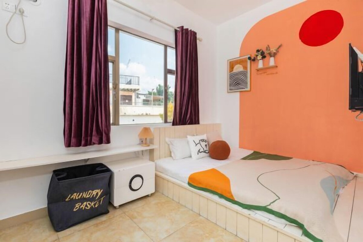 A bedroom with a bed, a window with curtains, a wall-mounted shelf with decorative items, and a laundry basket.
