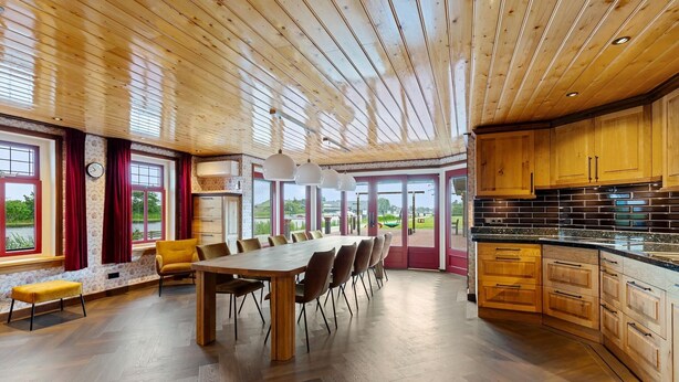 A spacious kitchen with wooden cabinets and a large island, a dining area with a long table and chairs, and large windows offering a view of the outdoors. - Detached Home in Friesland by Canal (Jistrum / Skûlenboarch)