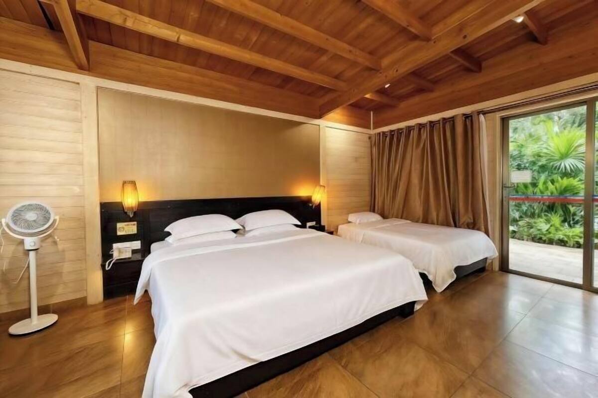 Ein Hotelzimmer mit zwei Betten, Holzbalkendecke, einem Standventilator und Blick ins Grüne durch das Fenster.