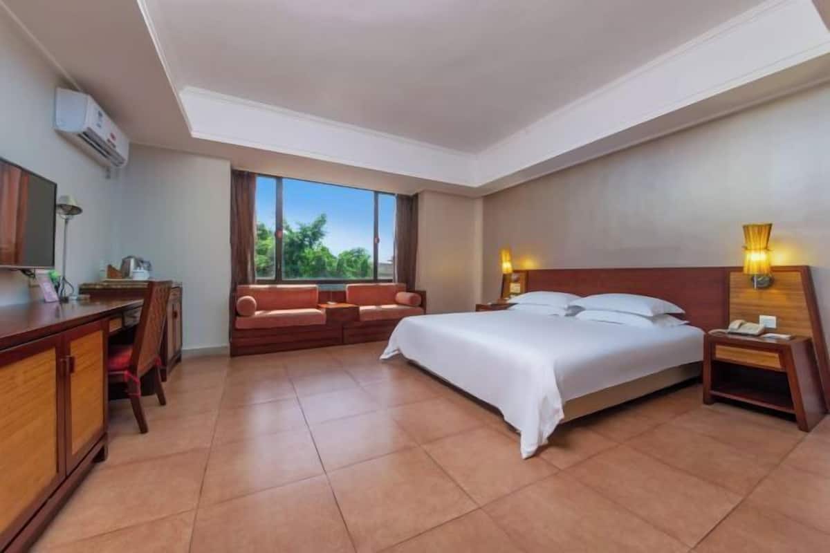 Ein Hotelzimmer mit Bett, Schreibtisch, Sessel, Sofa, Fernseher und einem Fenster mit Blick auf Grünflächen.