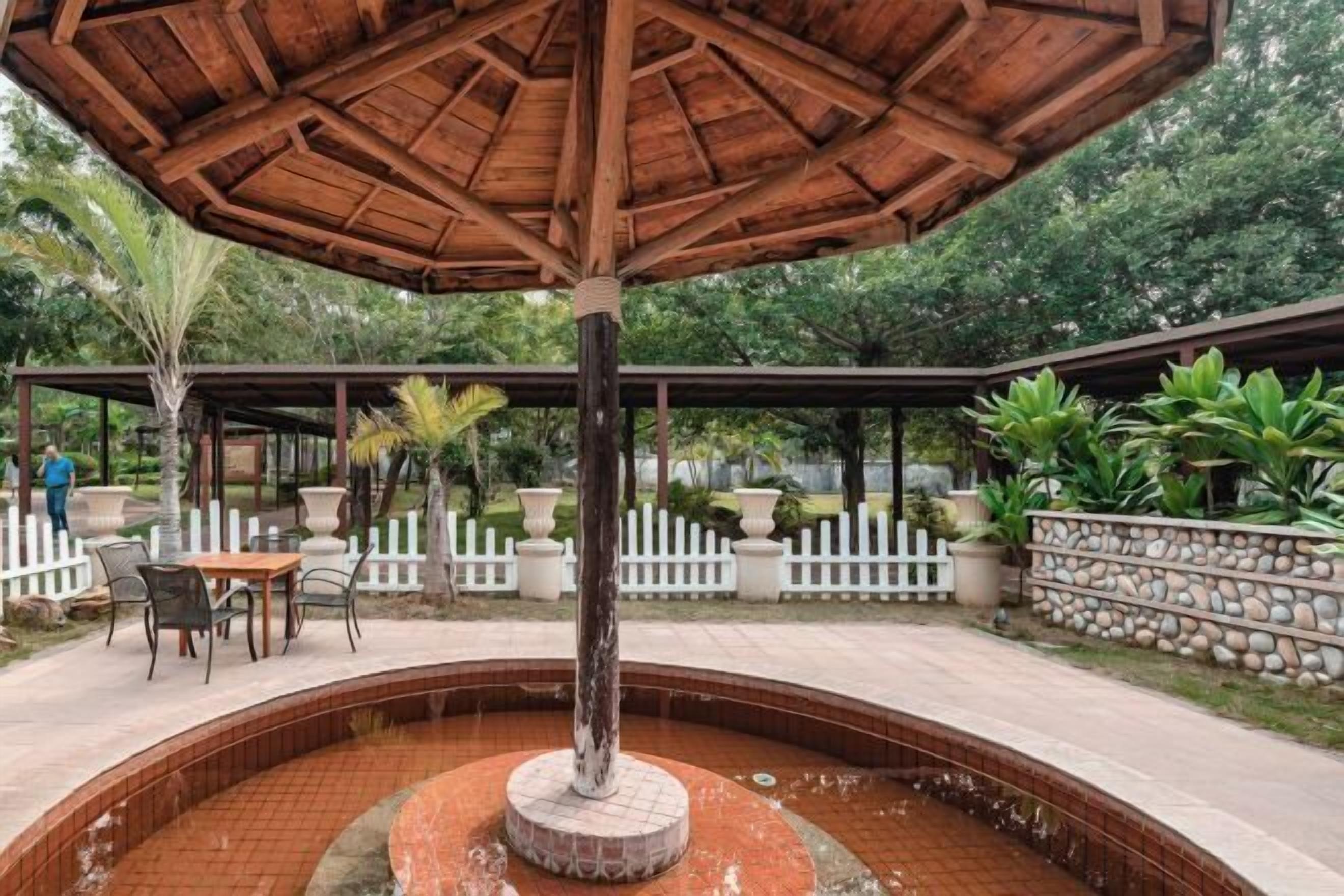 A covered outdoor area with a central fountain, surrounded by a white fence, potted plants, and a seating area with tables and chairs.