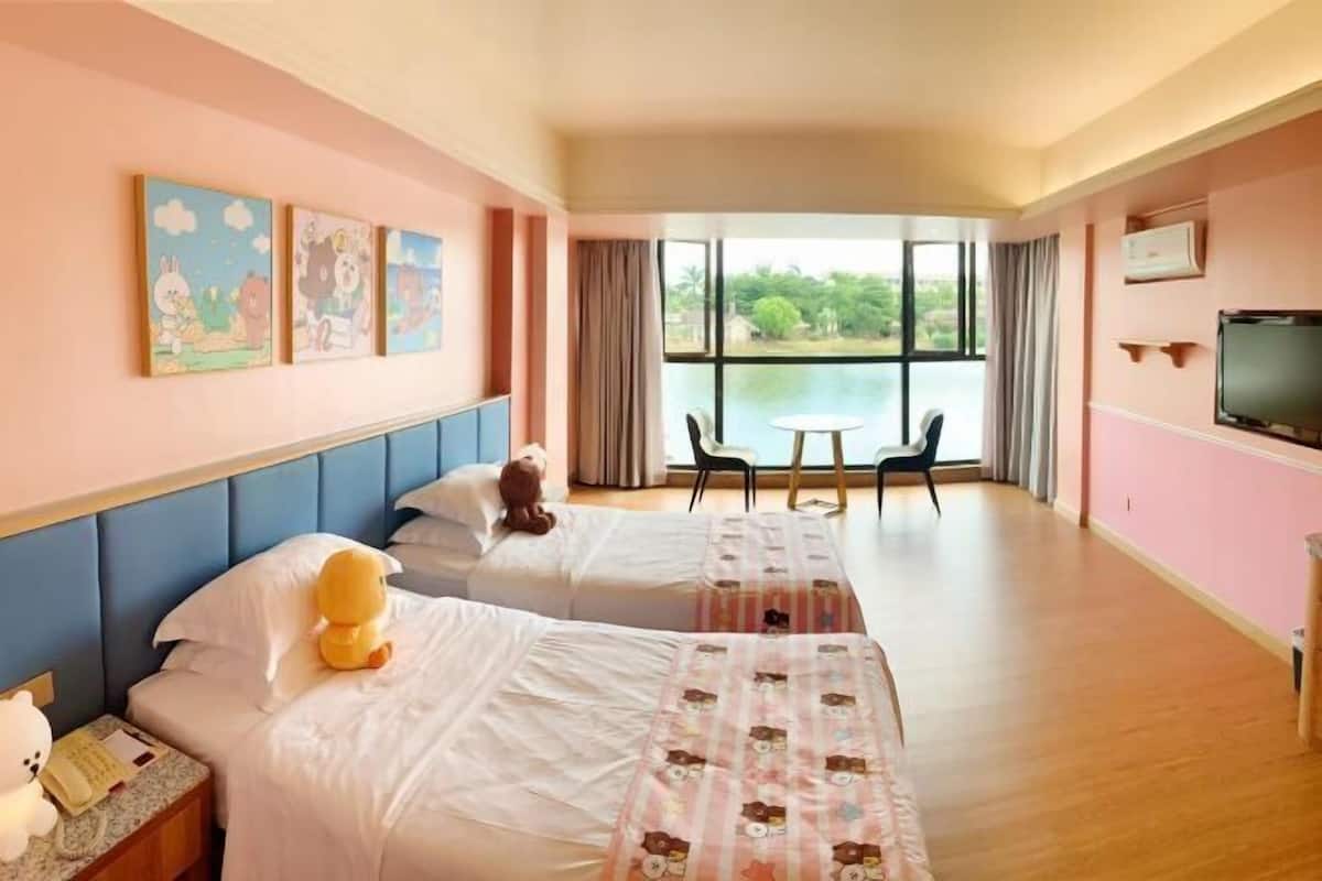 Ett hotellrum med två sängar, ett stort fönster med utsikt över grönområden, en TV och ett bord med stolar.