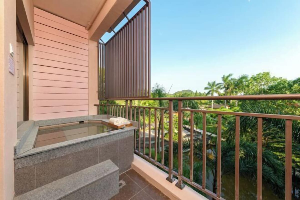 A balcony with a stone bench, a glass enclosure, and a view of a garden with palm trees.