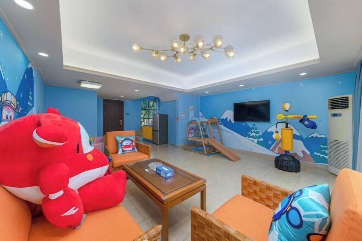 Ein Wohnzimmer mit einem großen, roten Plüschtier, orangefarbenen Möbeln, einem Holztisch, einem Fernseher und einem Wandbild mit Berglandschaft.