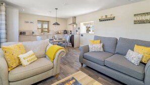 A modern living room with a grey sofa and a beige sofa, both with yellow pillows. There is a dining table with chairs, a kitchen area in the background, and a window with blinds. - Rosemary – Ground Floor (Boston)