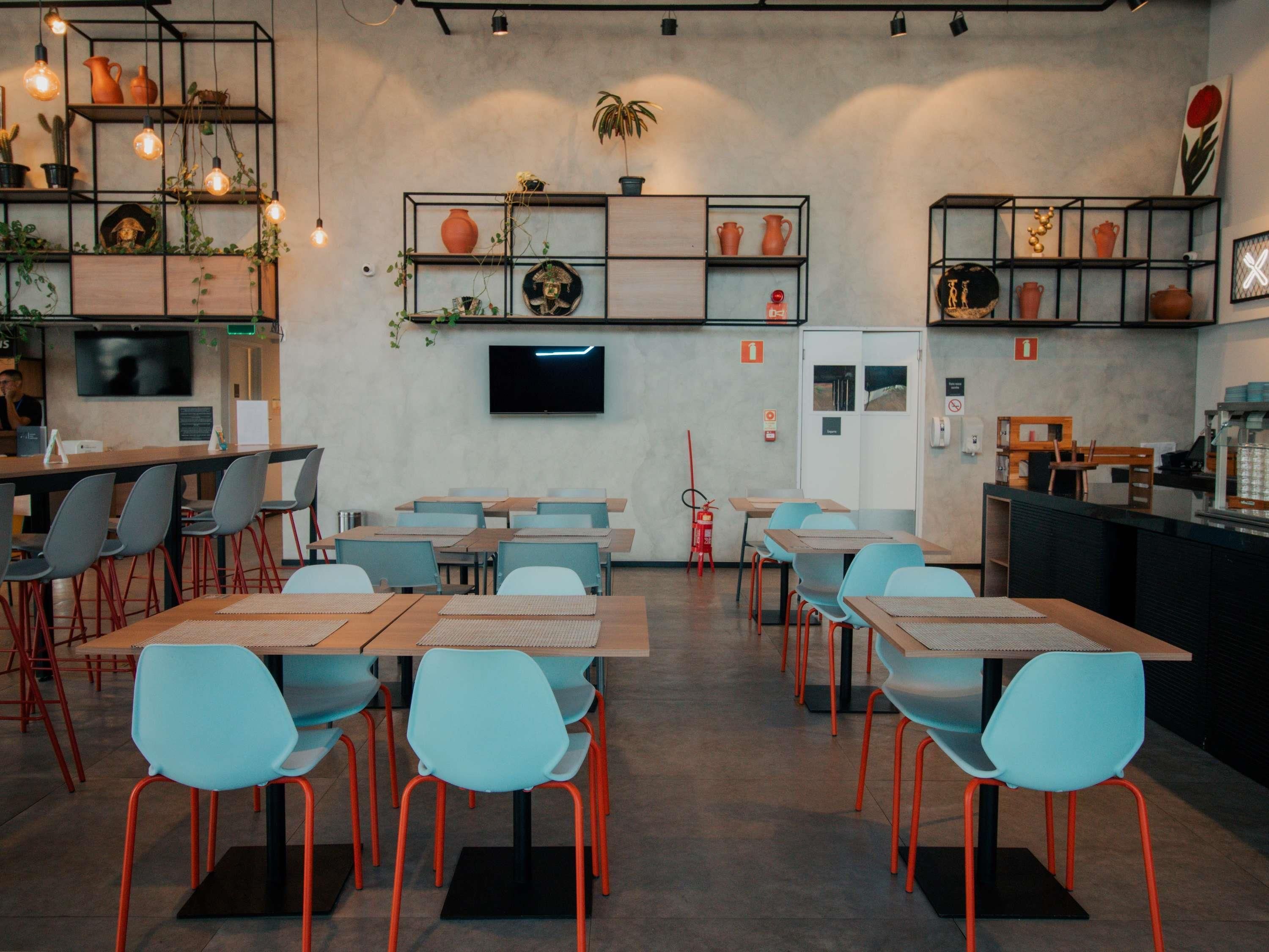 A modern cafe with multiple tables and chairs, a flat-screen TV on the wall, and shelves with plants and pottery.