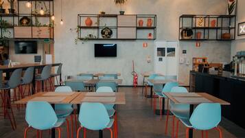 A modern cafe with multiple tables and chairs, a flat-screen TV on the wall, and shelves with plants and pottery.