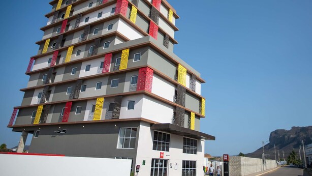 Un hôtel à plusieurs étages, doté d’une architecture moderne, avec des balcons ornés de garde-corps à motifs et une grande enseigne rouge bien visible sur le toit.