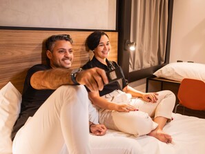 A couple sitting on a bed in a hotel room with a wooden headboard, a nightstand, and a red chair. - Ibis Serra Talhada (Serra Talhada)