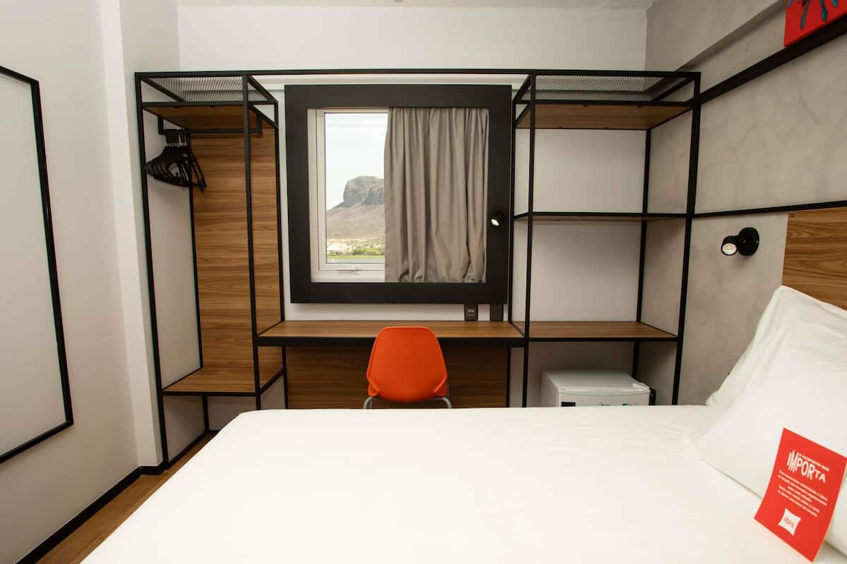 A compact hotel room with a bunk bed, a desk with a chair, a small bedside table, and a window with a view of a mountain.