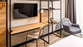 A modern hotel room with a wooden desk, a grey armchair, a TV mounted on the wall, and a shelf with a lamp and a coffee maker.