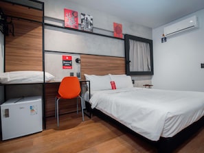 Hotellihuone, jossa on sänky, tuoli, pieni jääkaappi ja seinälle asennettu ilmastointilaite.