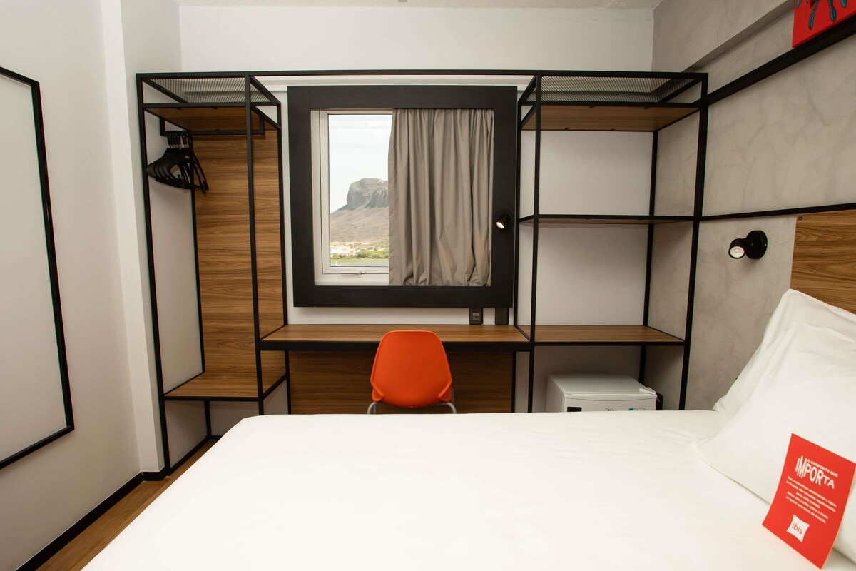 A compact hotel room with a bunk bed, a desk with a chair, a small bedside table, and a window with a view of a mountain.