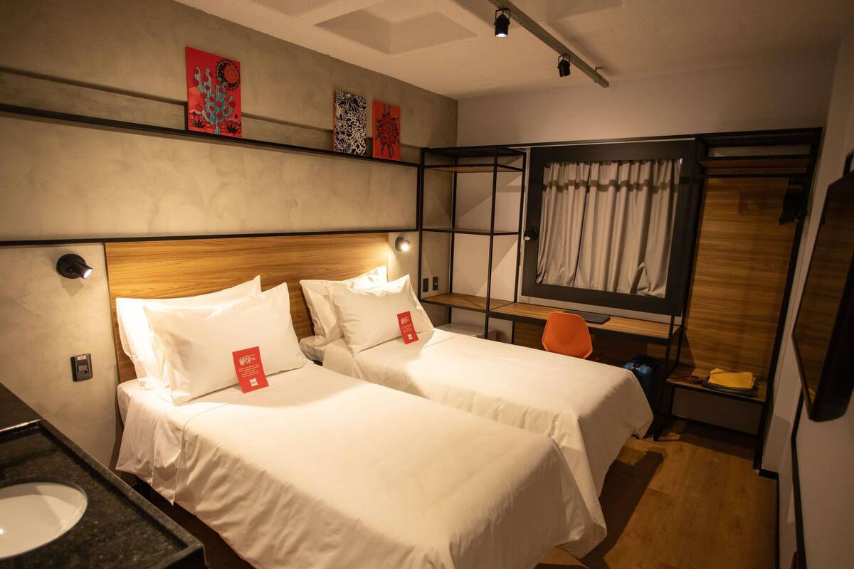 A hotel room with two beds, a desk, a chair, a mirror, and artwork on the wall.