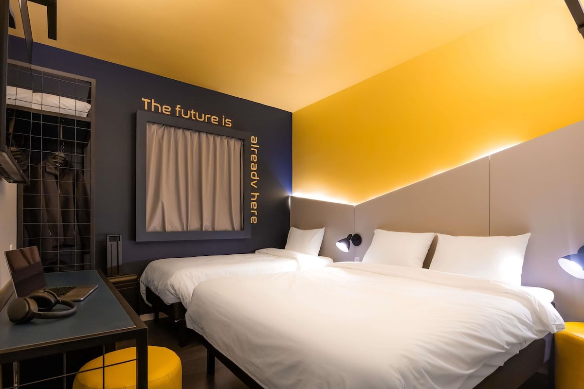 A modern hotel room with a large bed, a desk with a laptop, a chair, and a wardrobe.