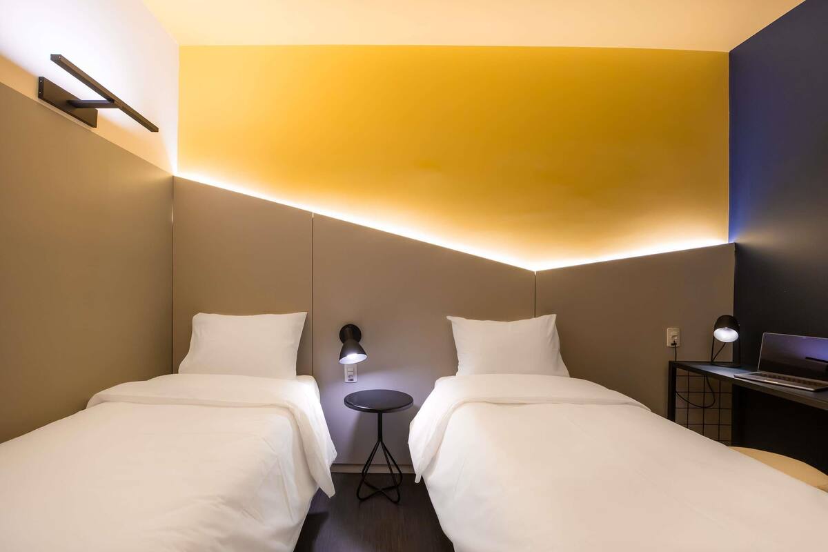 Ein Hotelzimmer mit zwei Betten, einem kleinen runden Tisch, einem Schreibtisch mit einem Laptop und einer Wandleuchte.