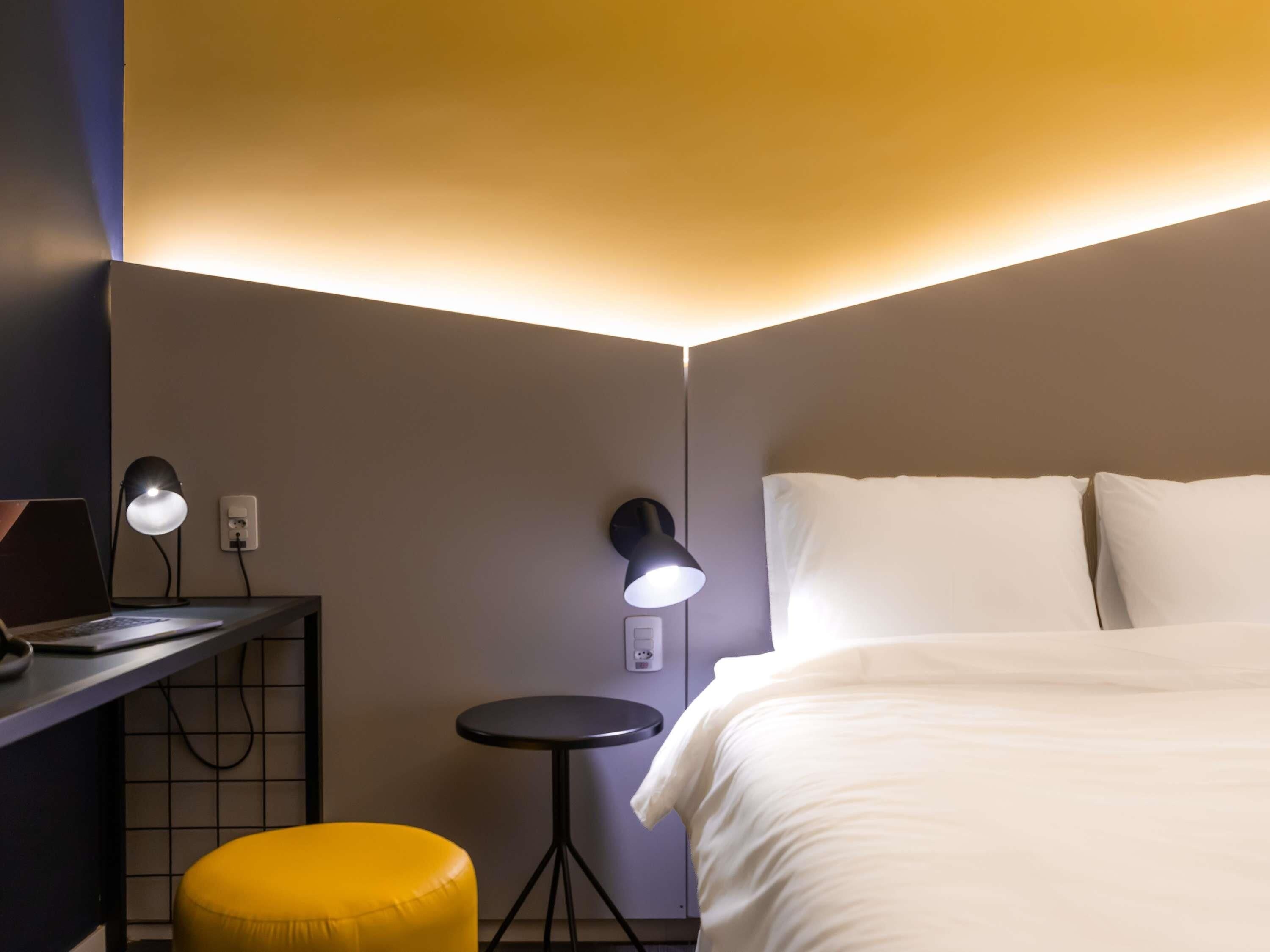 A hotel room with a bed, a desk with a laptop, a small table, and two wall-mounted lamps.