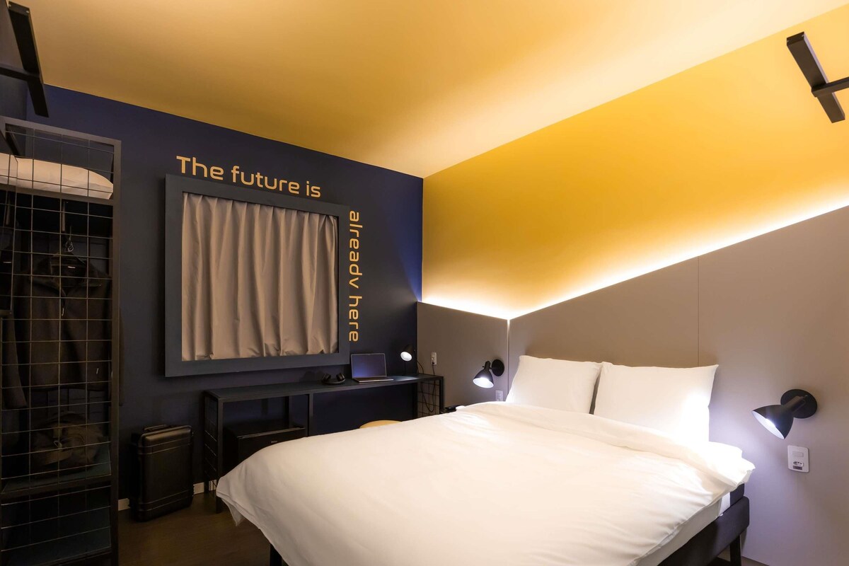 A modern hotel room with a bed, a desk, a chair, a wardrobe, and a mirror.